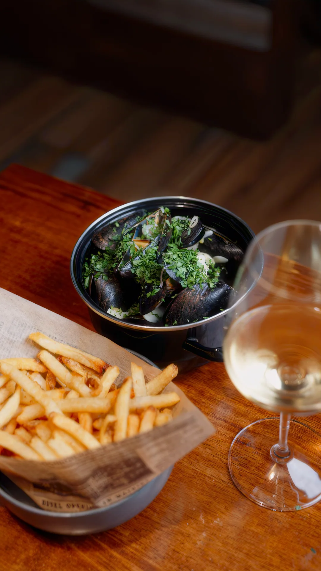 A copper pot of mussels scattered with parsley beside a paper cone of frites and a glass of white wine