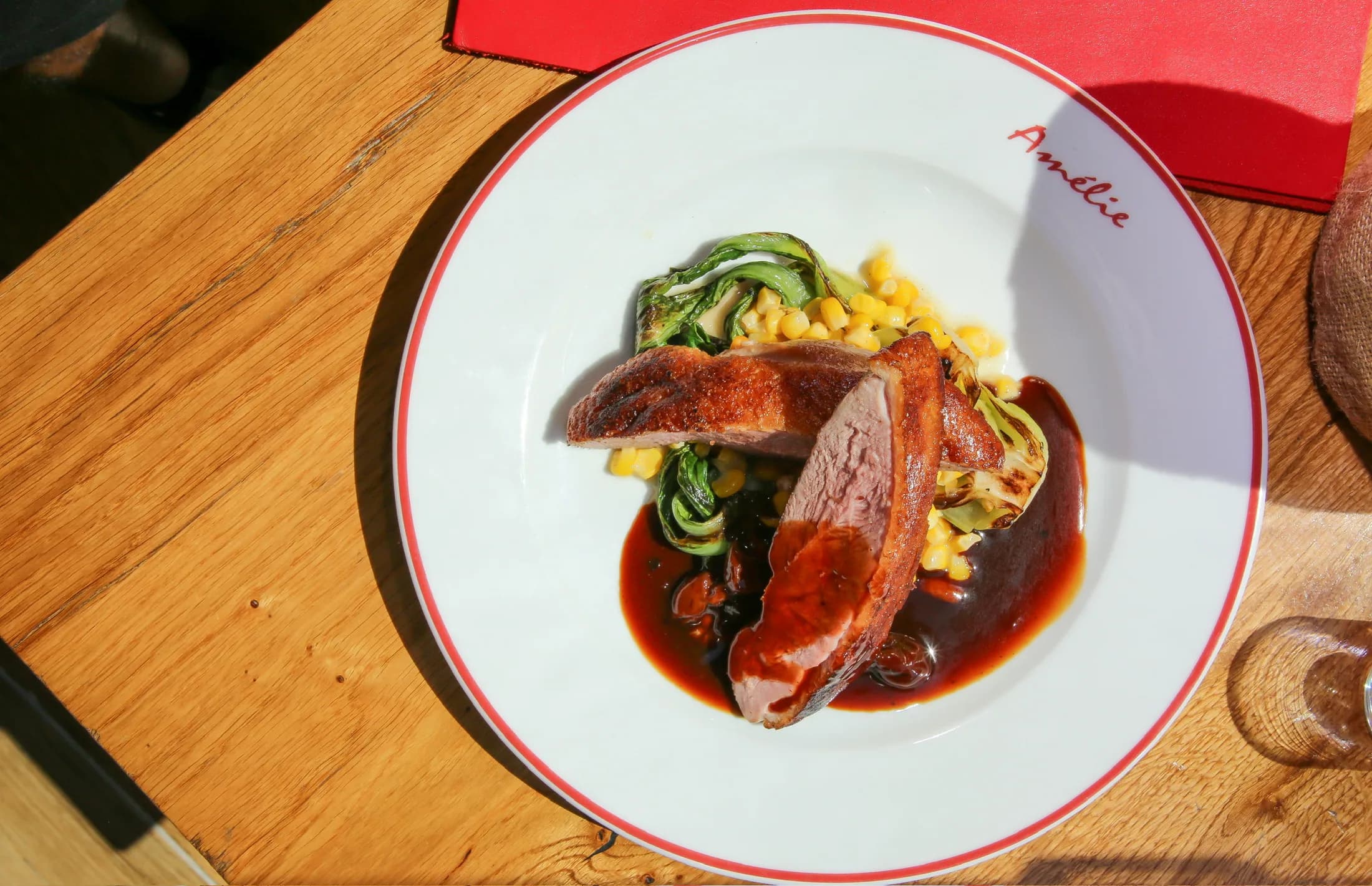 Sliced duck breast with corn and greens on an Amélie red-rimmed plate beside a red napkin, in hard afternoon sunlight