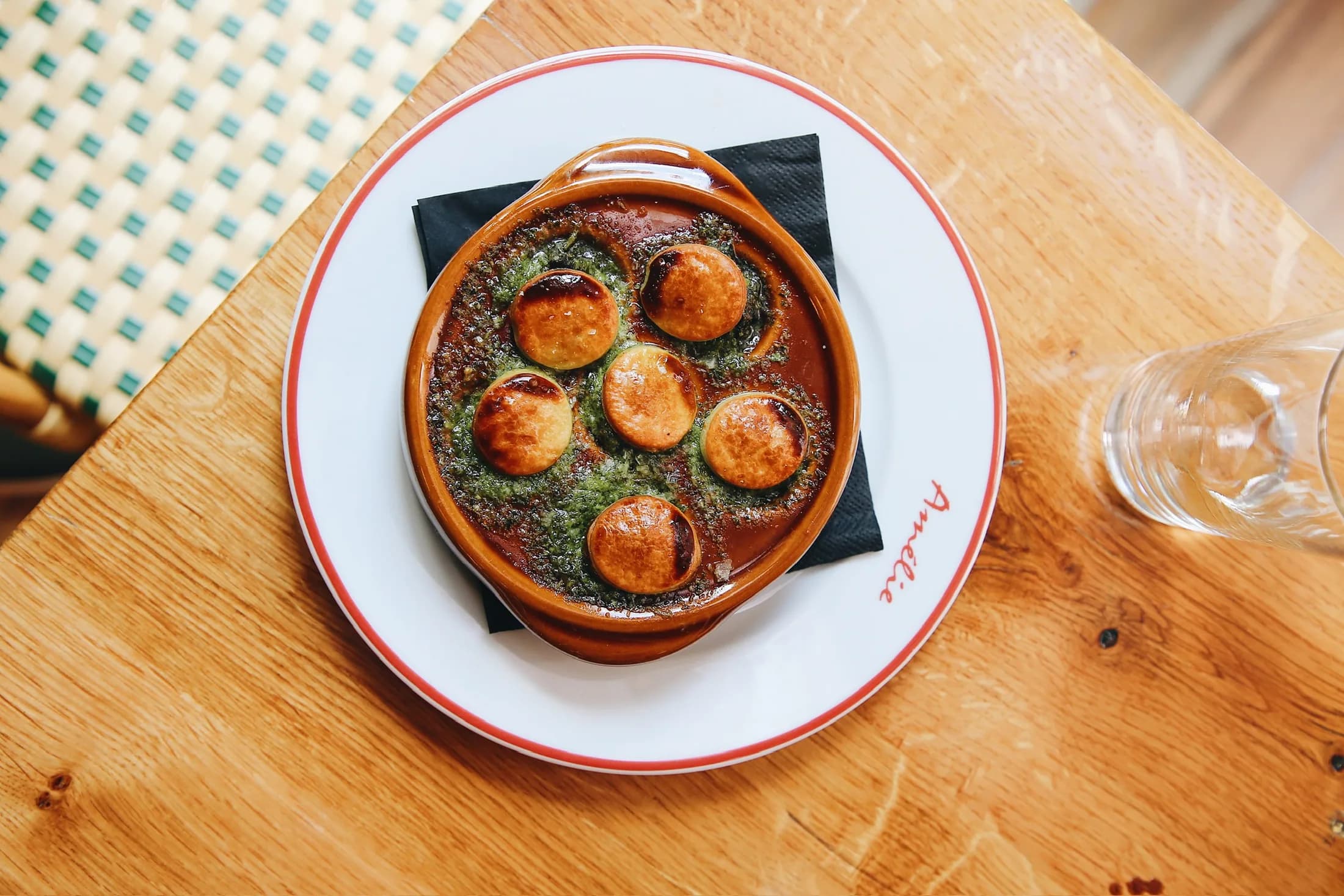 Escargots à l'ail in a terracotta dish on an Amélie red-rimmed plate, shot from above on a blond oak table