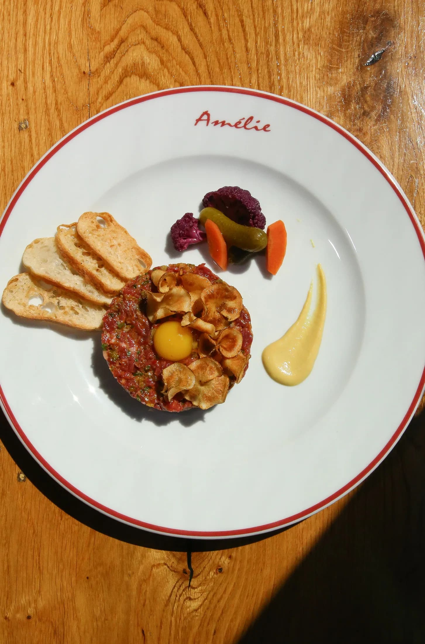 Hand-cut beef tartare with toast points and pickles on an Amélie red-rimmed plate, sunlight across the table